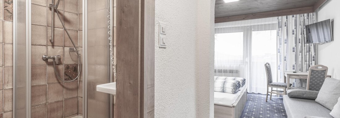 A modern bathroom with a shower and bright tiles. On the other side is a cozy living area with a sofa and a window. - Gästeheim Schmiedhof - Sölden