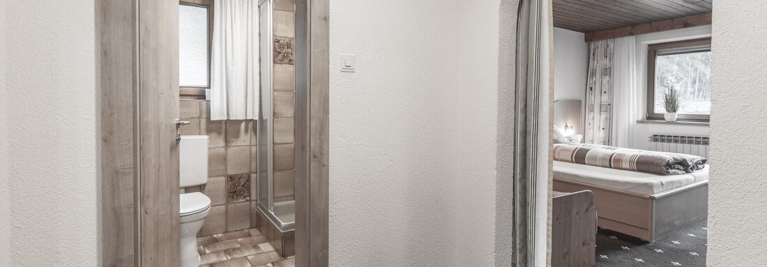 A bright room with an open passage to a bathroom. The walls are light, and the floor has a dark carpet with a simple pattern. - Gästeheim Schmiedhof - Sölden