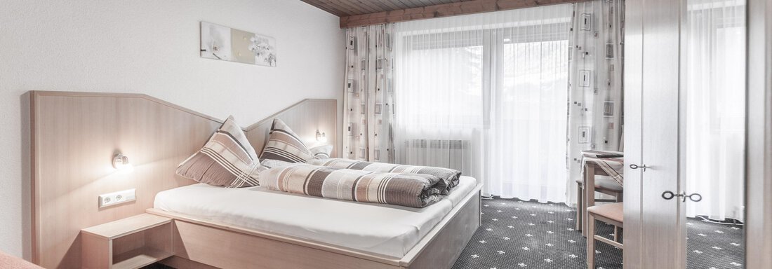 A cozy bedroom with two beds and wooden elements. Large windows let in plenty of light and offer a view outside. - Gästeheim Schmiedhof - Sölden