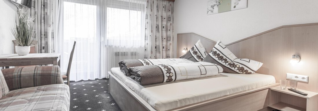 A cozy room with a double bed and a table. Large windows provide plenty of light and a beautiful view. - Gästeheim Schmiedhof - Sölden