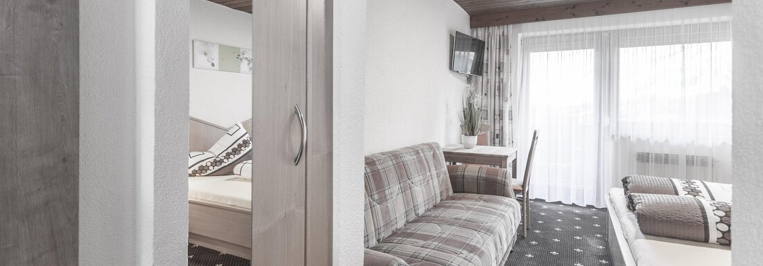 A modern living room with a gray sofa and a light wooden ceiling. Large windows provide plenty of natural light. - Gästeheim Schmiedhof - Sölden