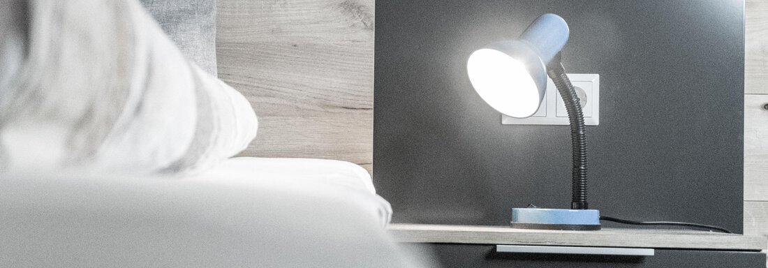 A modern table lamp shines on a nightstand. Wooden paneling is visible in the background. - Gästeheim Schmiedhof - Sölden