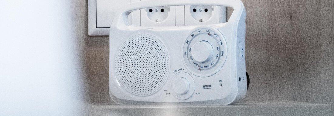 A white radio stands on a table. In the background, power outlets are visible. - Gästeheim Schmiedhof - Sölden