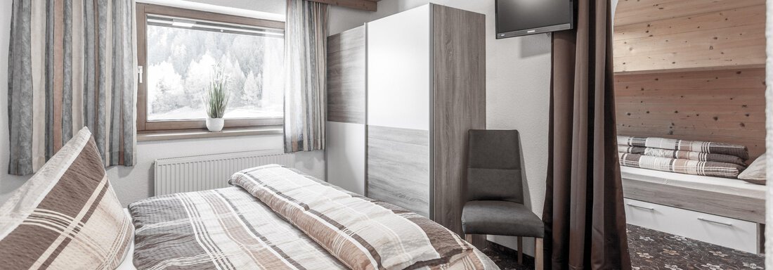 A cozy bedroom with a large bed and gray walls. Next to the bed is a comfortable chair and a small television on the wall. - Gästeheim Schmiedhof - Sölden