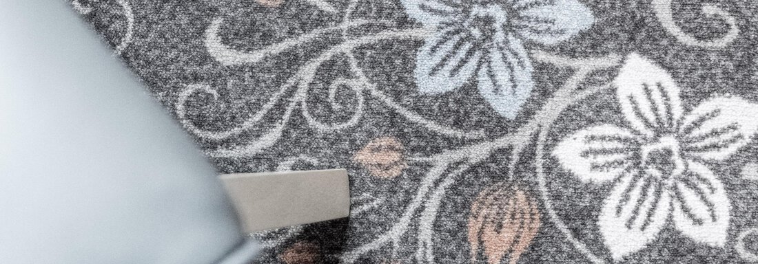A carpet with floral patterns in gray, blue, and soft colors. On the edge, a chair is visible, complementing the design. - Gästeheim Schmiedhof - Sölden