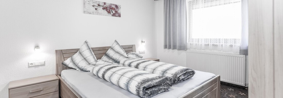 A cozy bedroom with a large bed and striped blankets. Bright walls and windows create a friendly atmosphere. - Gästeheim Schmiedhof - Sölden