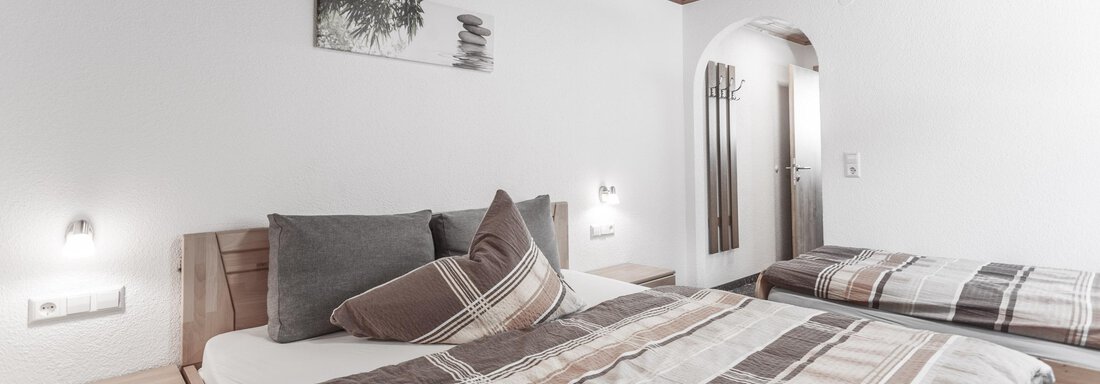 A cozy bedroom with a large bed and a single bed. The walls are white, and there are wooden details as well as soft lighting. - Gästeheim Schmiedhof - Sölden