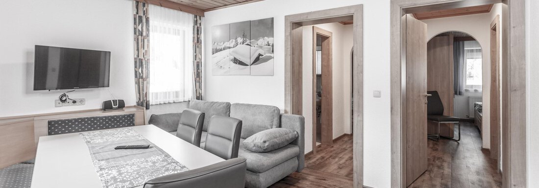 A modern living room with a sofa and a dining table. The walls are light, and there is a television as well as a passage to other rooms. - Gästeheim Schmiedhof - Sölden