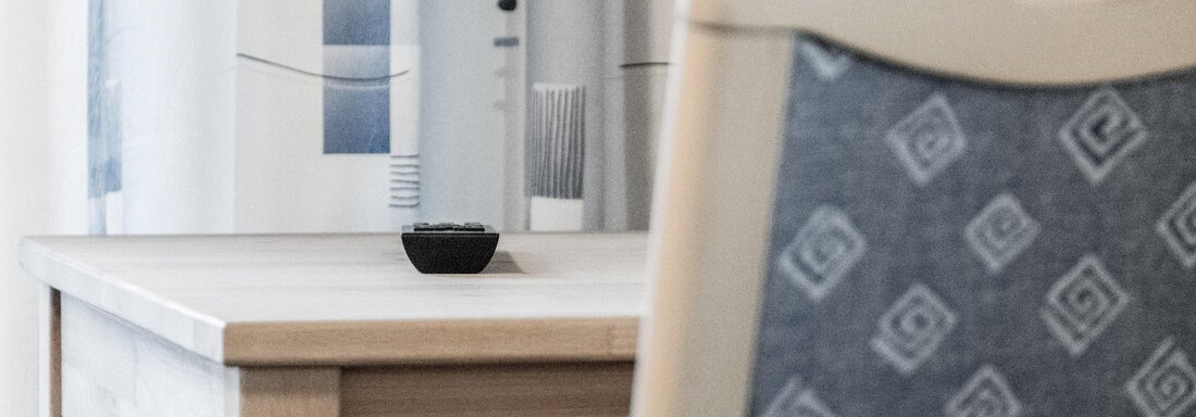 A simple table with a small black object on it. In the background, a chair and curtains are visible. - Gästeheim Schmiedhof - Sölden
