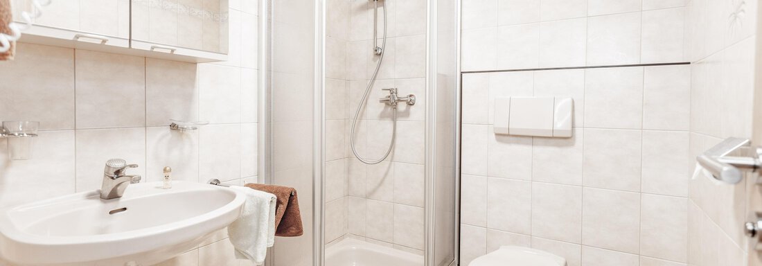 A modern bathroom with a shower, a sink, and a toilet. The walls are brightly tiled, and the furnishings are simple and functional. - Gästeheim Schmiedhof - Sölden