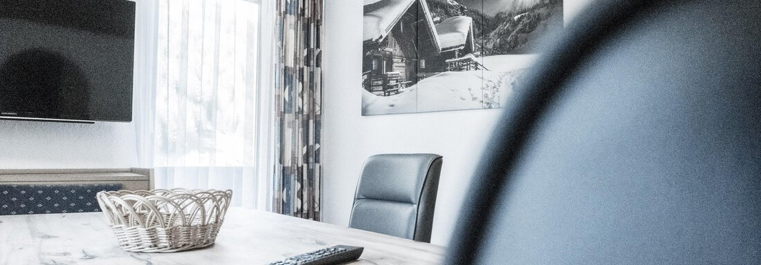A modern furnished room with wooden decor and a large table. On the wall hangs a picture of a winter mountain house. - Gästeheim Schmiedhof - Sölden