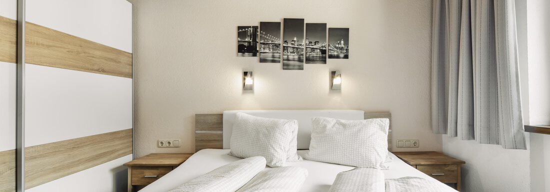 A modern bedroom with a double bed and a wooden ceiling. On the wall hangs a picture with a city view, and there are bright curtains. - Gasthof Granstein - Sölden