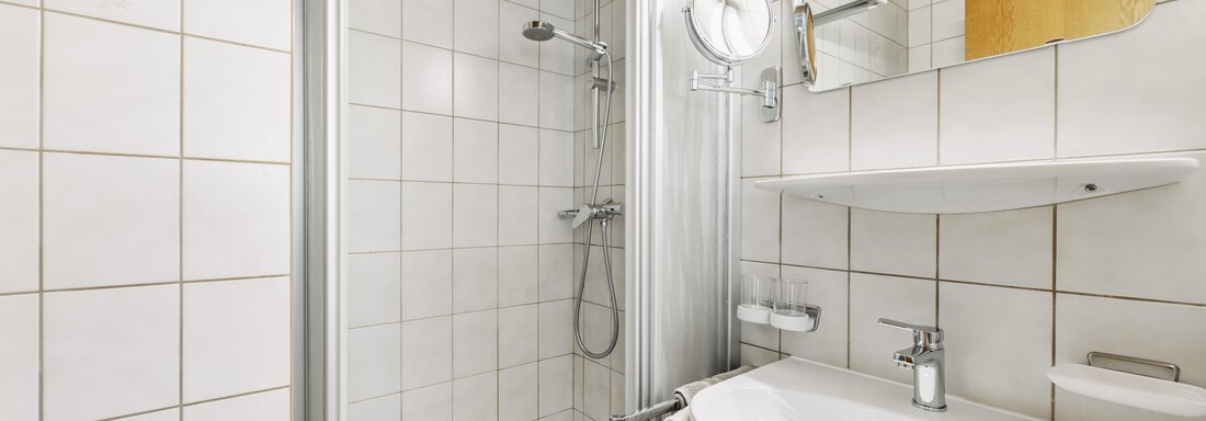 A modern bathroom with a shower cabin and a sink. The walls are tiled with white tiles and there is a mirror above the sink. - Gasthof Granstein - Sölden