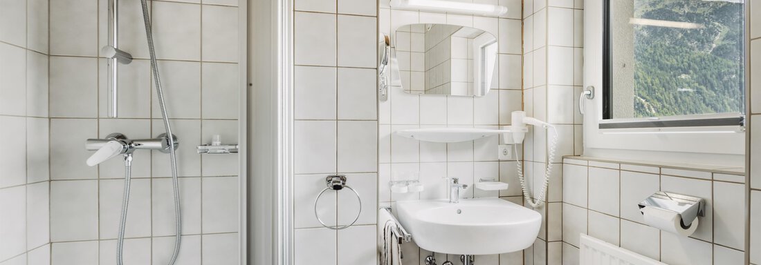 A modern bathroom with white tiles and a shower. There is a sink, a mirror, and a window with a view. - Gasthof Granstein - Sölden