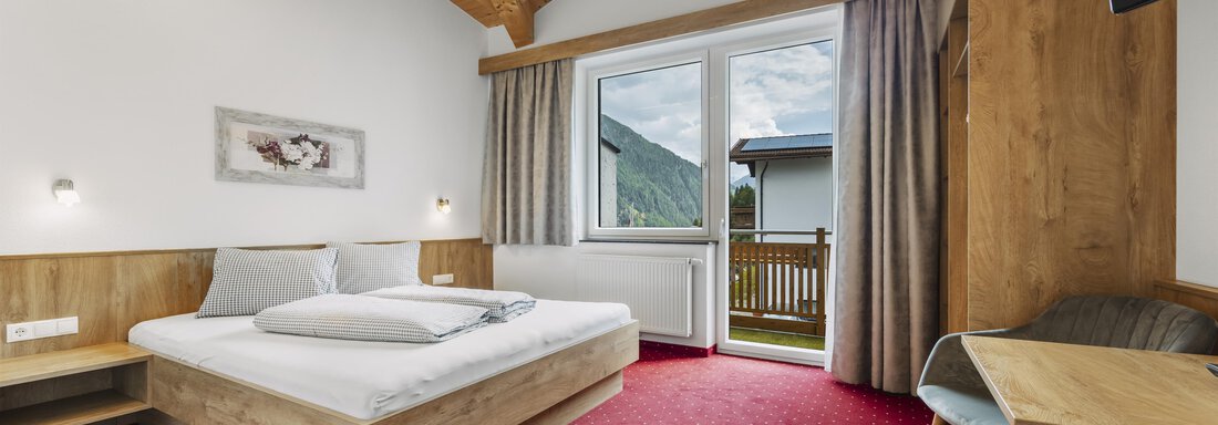 A bright room with a wooden ceiling and a red carpet. It contains a bed, a table, and a balcony with a view outside. - Gasthof Granstein - Sölden