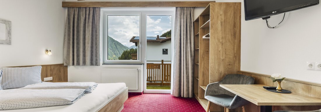 A bright hotel room with wooden furniture and a large window. There is a bed, a table, and a television. - Gasthof Granstein - Sölden