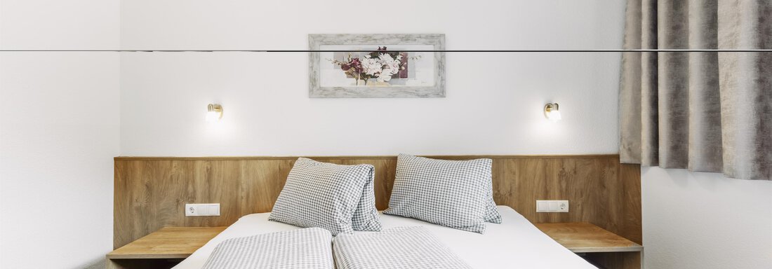 A modern bedroom with two single beds and gray pillows. The walls are plain and there is a curtain as well as wall lamps. - Gasthof Granstein - Sölden