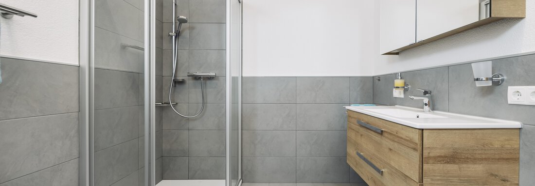 A modern bathroom with a clear shower cabin and a wooden washbasin. The walls are in neutral gray tones, giving it an elegant look. - Gasthof Granstein - Sölden