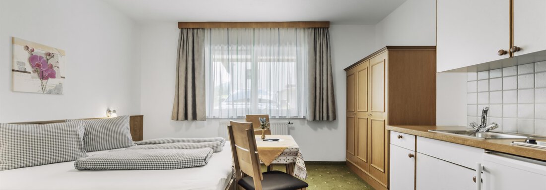 A simple room with two single beds and a small kitchen area. Large windows let in plenty of light and provide a bright atmosphere. - Gasthof Granstein - Sölden