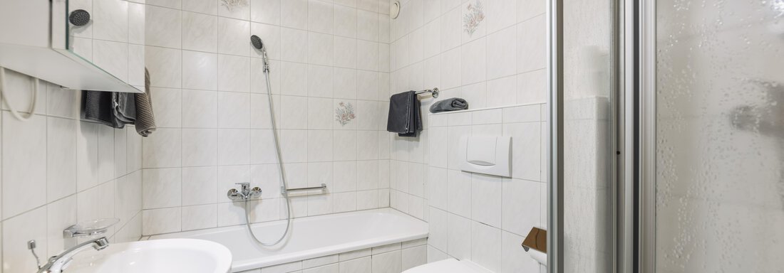 A modern bathroom with a shower, bathtub, and sink. The walls are tiled, and there are wood panelings on the ceiling. - Gasthof Granstein - Sölden