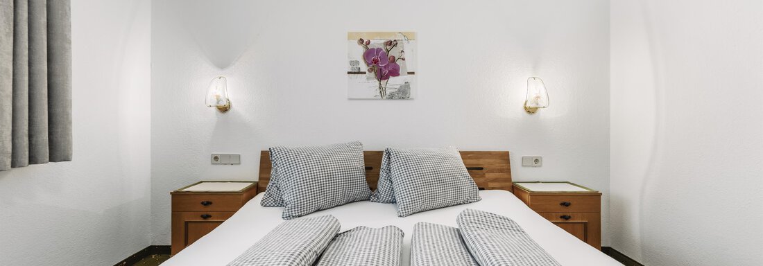 A cozy bedroom with a large bed and striped pillows. On the walls hang lamps and a small piece of art. - Gasthof Granstein - Sölden