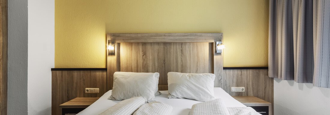 A cozy bedroom with a double bed and two pillows. The walls are painted yellow and the windows are decorated with gray curtains. - Gasthof Granstein - Sölden