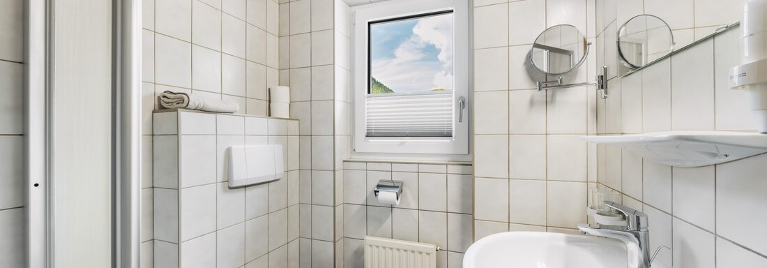 A modern bathroom with white tiles and a wooden ceiling. It features a sink, a toilet, and a window with a view. - Gasthof Granstein - Sölden