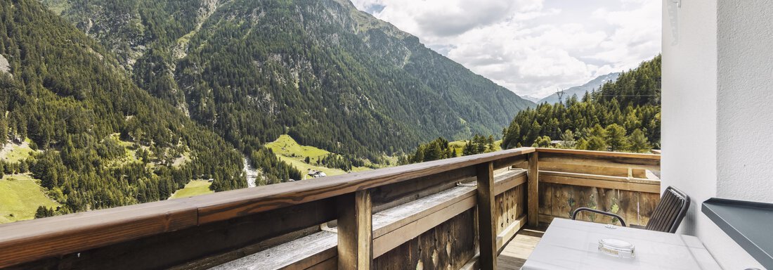 A balcony with a wooden railing and two chairs offers a stunning view of the mountains. The sky is cloudy and the landscape is green and hilly. - Gasthof Granstein - Sölden