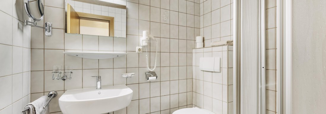A modern bathroom with white tiles and a shower cubicle. It contains a sink, a toilet, and a mirror. - Gasthof Granstein - Sölden