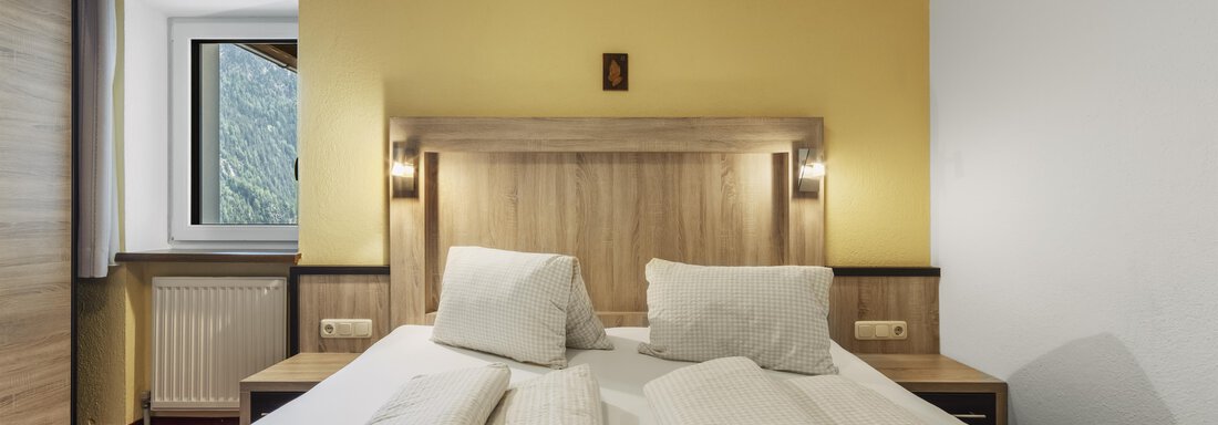 A cozy bedroom with a double bed and light wooden interior. The walls are painted in a warm yellow, and a window lets in plenty of daylight. - Gasthof Granstein - Sölden