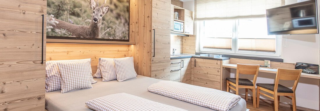 A cozy wooden room with a double bed and checkered pillows. In the background, a picture of a deer and a small kitchenette are visible. - GIGGI Tenne Studios - Sölden