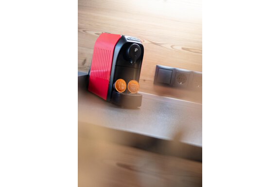 A red coffee machine is on a table. Next to the machine are two coffee capsules. - GIGGI Tenne Studios - Sölden