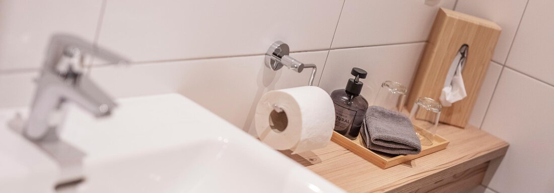 A modern bathroom with an elegant sink and a wooden shelf. On the shelf are care products, towels, and toilet paper. - GIGGI Tenne Studios - Sölden