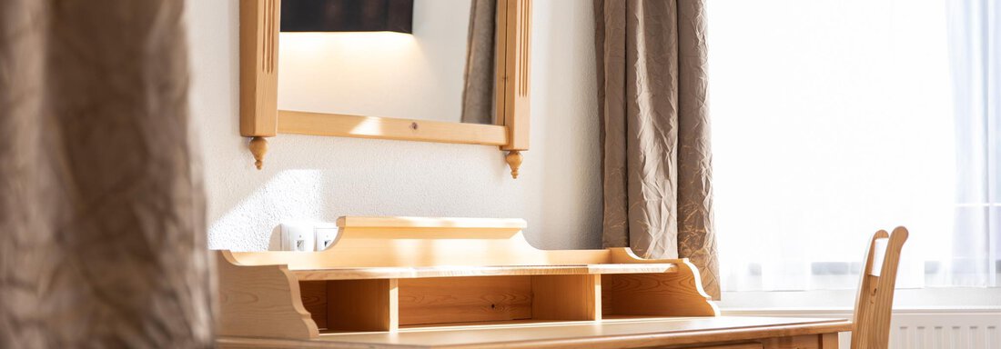 Ein heller und gemütlicher Schreibtisch aus Holz mit einem Spiegel darüber. Das Fenster lässt viel Tageslicht herein und die Vorhänge sind in sanften Brauntönen gehalten. - Glanz & Glory Hotel - Sölden