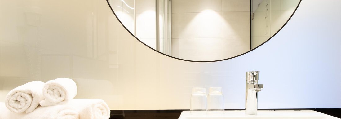 Ein modernes Badezimmer mit einem quadratischen Waschbecken und einem großen runden Spiegel. Saubere Handtücher liegen auf einem Holzregal. - Glanz & Glory Hotel - Sölden