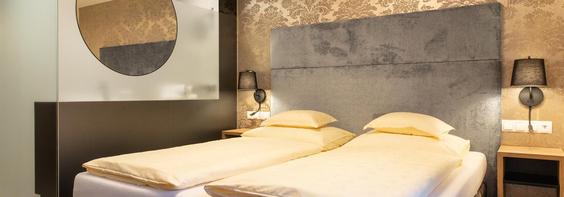 Ein modernes Zimmer mit zwei Einzelbetten und stilvoller Dekoration. Ein elegantes Badezimmer mit einer runden Wandspiegel-Elemente ist ebenfalls sichtbar. - Glanz & Glory Hotel - Sölden