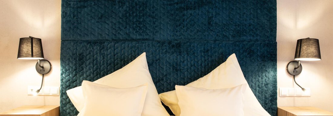 Ein gemütliches Schlafzimmer mit einem großen, gepolsterten Kopfteil in Blau. Auf beiden Seiten des Bettes befinden sich elegante Nachttische mit Lampen. - Glanz & Glory Hotel - Sölden
