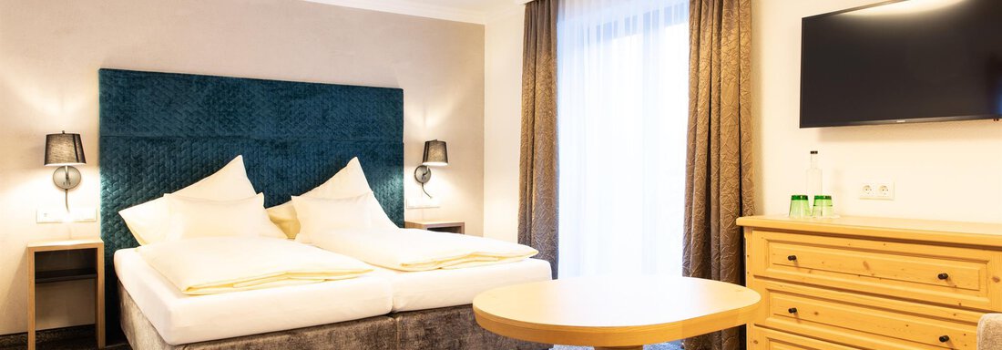 Ein modernes Hotelzimmer mit zwei Betten und einem runden Tisch. Der Raum ist hell und freundlich mit einem großen Fenster und einem Fernseher an der Wand. - Glanz & Glory Hotel - Sölden