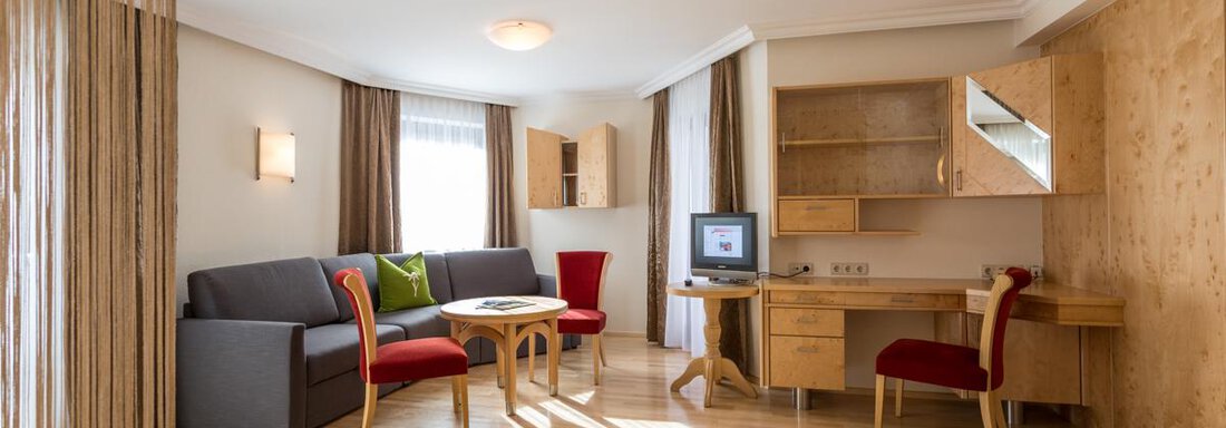 Wohnraum Junior Suite Nederkogel | © Hotel Stefan (CC C) Ein helle und freundliche Wohnzimmer mit einem grauen Sofa, einem Tisch und zwei roten Stühlen. An der Wand befindet sich ein Holzregal und ein Fernseher. - Glanz & Glory Hotel - Sölden