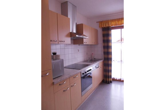 A modern kitchen with light wood cabinets and a window with gingham-patterned curtains. The equipment includes a stove, a sink, and a table with chairs. - Gstrein Andreas - Sölden