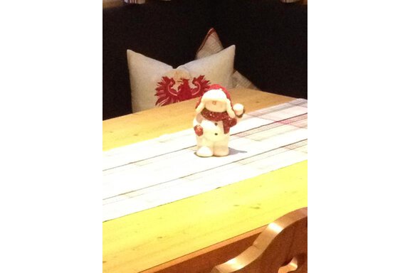 A decorative snowman figure stands on a wooden table with a table runner. In the background, a cushion with a red pattern can be seen. - Haus Almrausch - Sölden