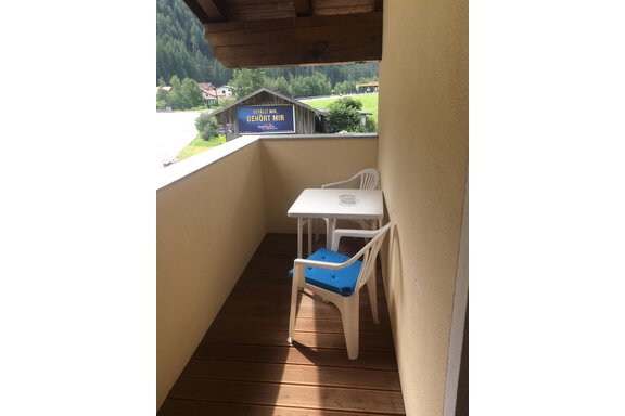 A small balcony with a table and two chairs. In the background, trees and a wooden building are visible. - Haus Almrausch - Sölden