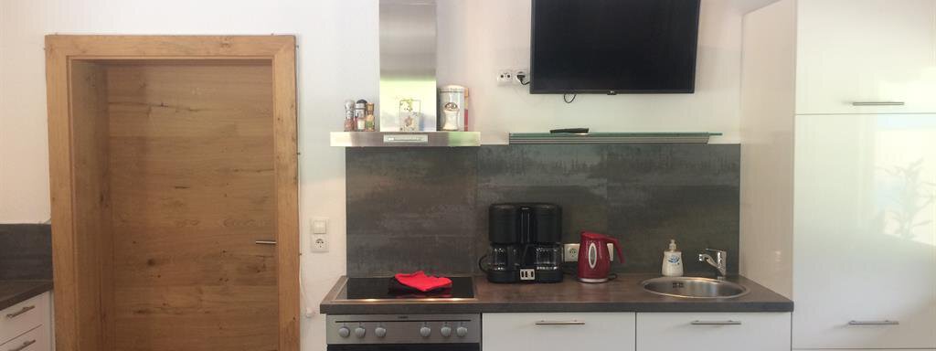 A modern kitchen with white cabinets, a stove, and a sink. On the wall, there is a television and a door leading to another room. - Haus Almrausch - Sölden