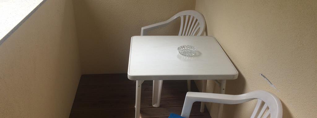 A small balcony with two white chairs and a table. On the table, there is an ashtray, and in the background, green meadows are visible. - Haus Almrausch - Sölden