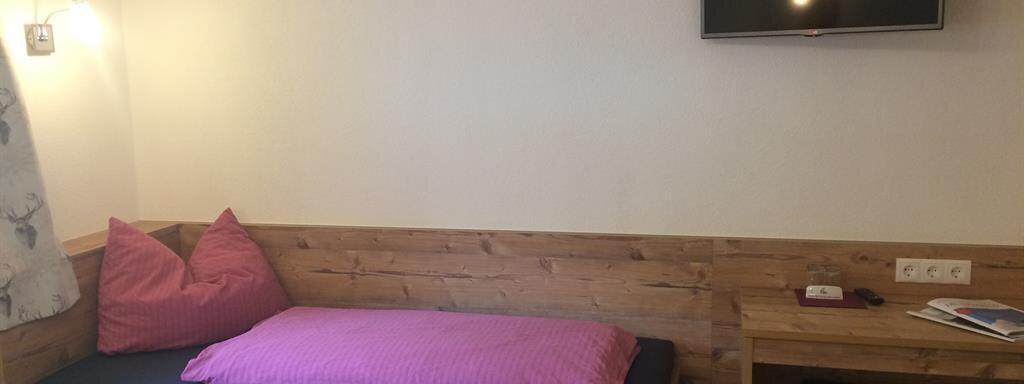 A cozy room with a single bed, wooden paneling, and a television on the wall. There is also a small table and a chair next to the bed. - Haus Almrausch - Sölden