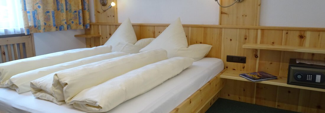 A cozy bedroom with a wooden bed and two large pillows. The walls are paneled with wood, and the windows are decorated with colorful curtains. - Haus Alpenstern - Sölden