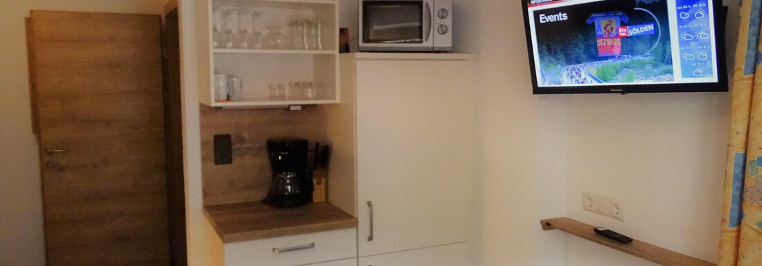 A small kitchenette with a refrigerator, microwave, and coffee maker. A television hangs on the wall displaying current information. - Haus Alpenstern - Sölden