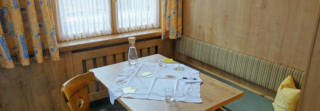 A cozy dining area with a wooden table and chairs. Curtains hang at the windows, and on the table are napkins and a water carafe. - Haus Alpenstern - Sölden