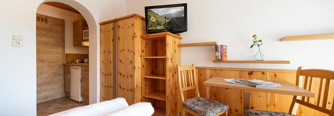 A cozy living space with wooden furniture and a TV on the wall. The room features a small dining table and a view of the kitchen. - Haus Alpenstern - Sölden
