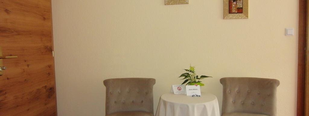 A cozy room with two beige chairs and a small table.  
On the wall hang two pictures, and on the table is a plant and a card. - Haus Amaris - Sölden